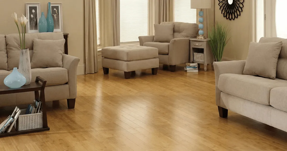 A living room with professionally installed bamboo flooring, beige sofas, a matching ottoman, a side table with a blue lamp, vases, framed pictures, and large windows with blinds and curtains in Omaha, NE